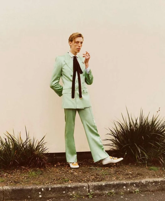 A blond-haired person in a mint green suit poses outdoors against a light wall near shrubs, wearing dress shoes and a black necktie.