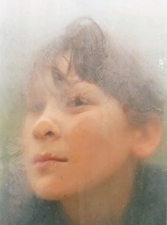 Close-up portrait of a young child looking up with a gentle expression, framed by a diffused and slightly obscured view.