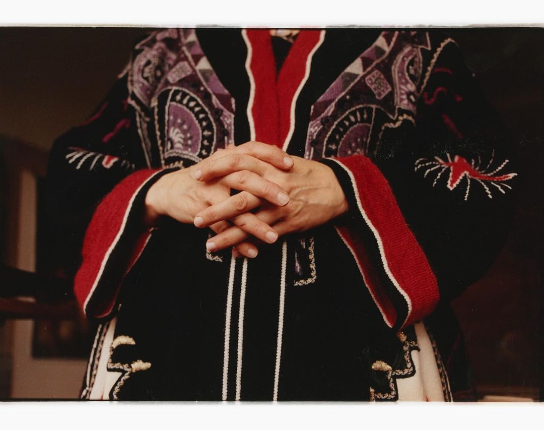 Person's hands folded over a black patterned folk-style sweater with red and white details, in an indoor setting with dark background.