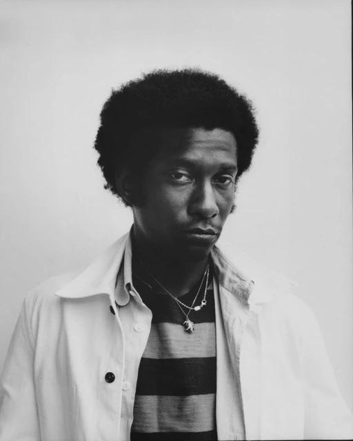 Portrait of a young Black man with an afro, wearing a striped shirt and a white jacket against a plain gray background.
