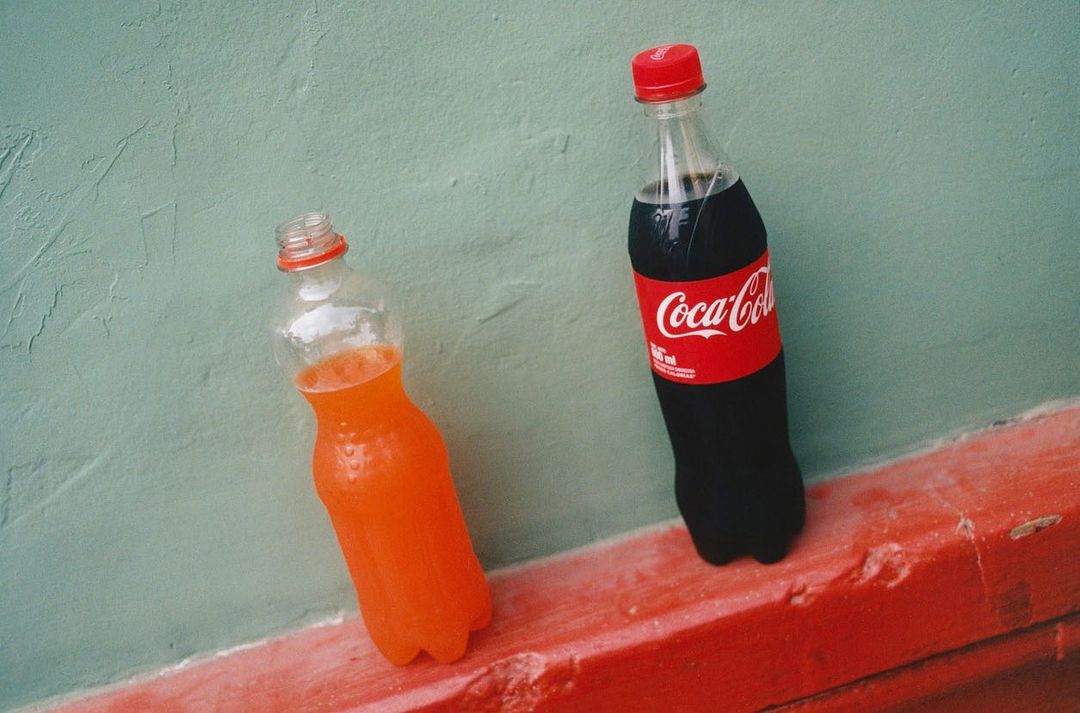 Two bottles of soda, one Coca-Cola and one orange soda, stand side-by-side against a light green wall and red trim for refreshing drinks.