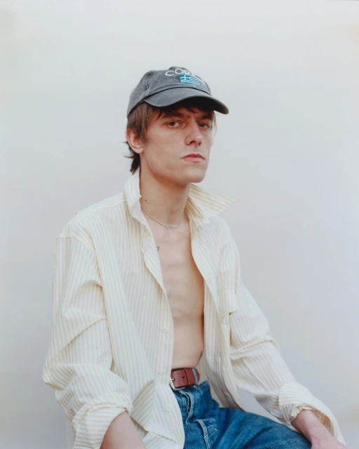 Young adult male poses indoors wearing a baseball cap, striped shirt, blue jeans, and brown belt against a white backdrop.