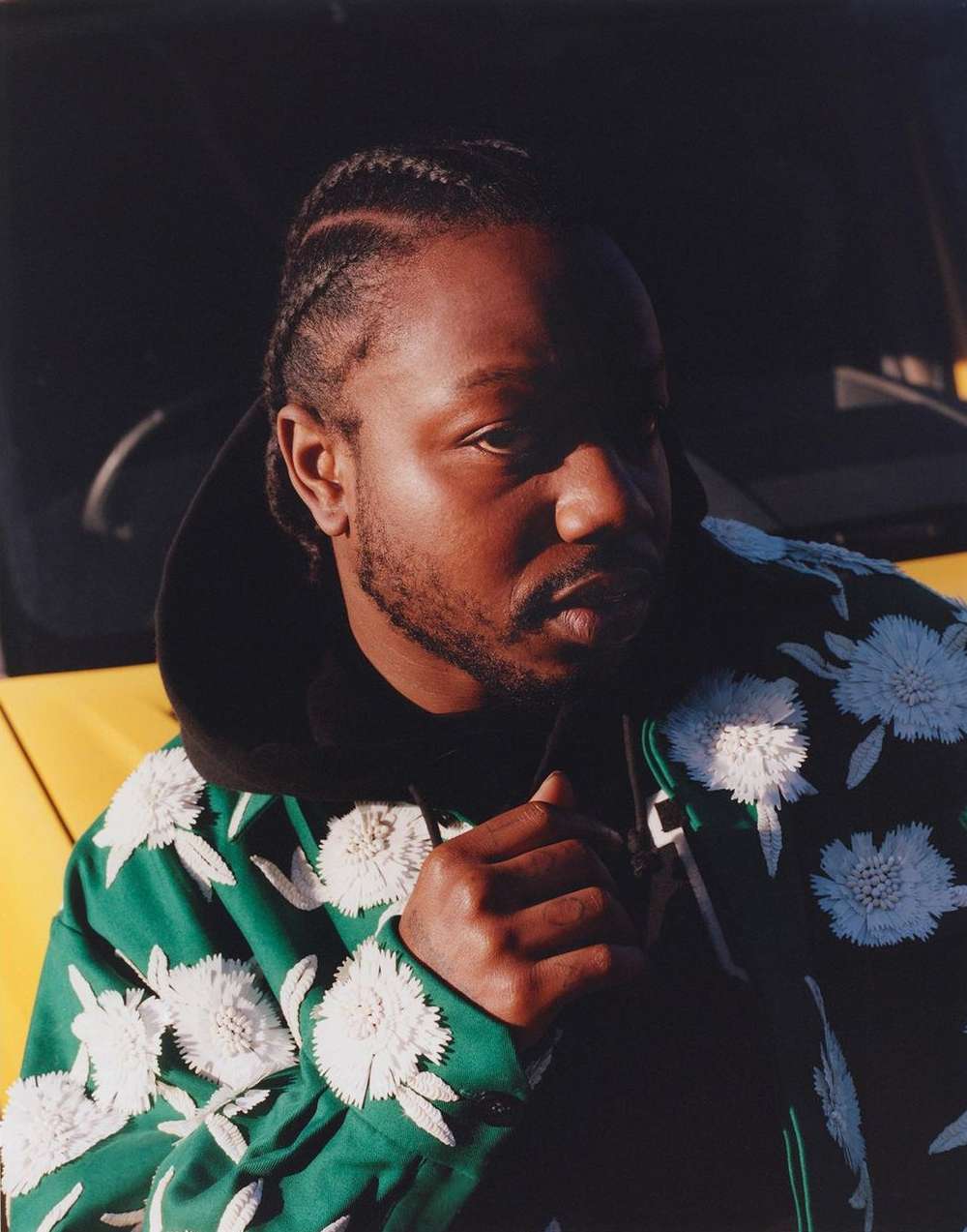 Portrait of an African-American man in a floral jacket, gazing to the side, with a yellow car partially visible in the background.