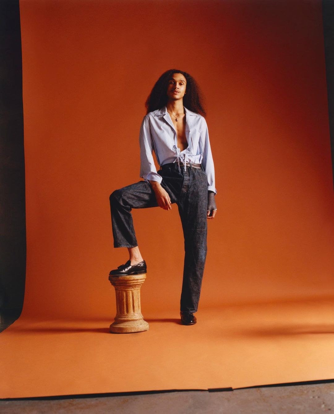 Full-length shot of a young adult female with dark hair posing with one foot on a plinth, set against an orange backdrop.