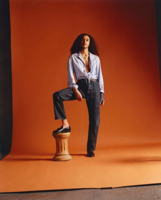 Full-length shot of a young adult female with dark hair posing with one foot on a plinth, set against an orange backdrop.
