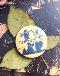 Round yellow Knebworth pin badge resting on top of three dried autumn leaves against a dark patterned surface.