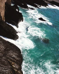 High angle photograph of turquoise ocean waves crashing against dark rocky coastal cliffs with white sea foam.
