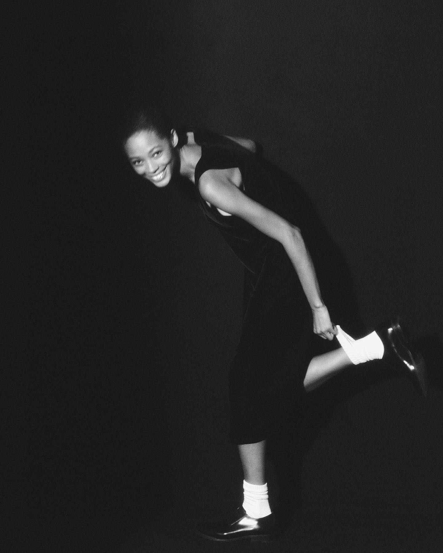 Woman in black dress smiles, extends leg to pull up a white sock in a studio with black background. Fashionable pose in monochrome.