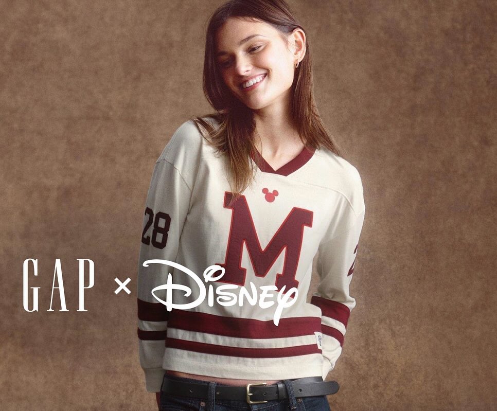 Smiling young woman poses in a vintage-style long-sleeved top with a Disney and Gap logo on a mottled brown background.