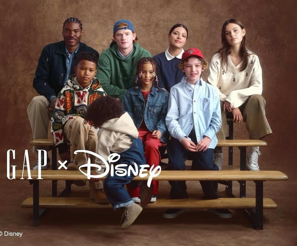 Group portrait with diverse children and adults in casual attire on bleachers, showcasing a collaborative GAP x Disney collection, on a brown background.