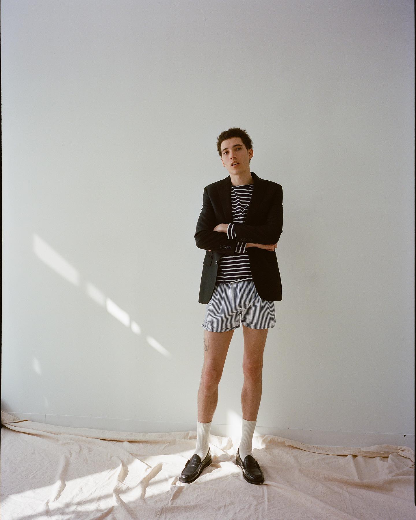 A young adult man in shorts, loafers, and a blazer poses indoors against a neutral backdrop with soft lighting and his arms are crossed.