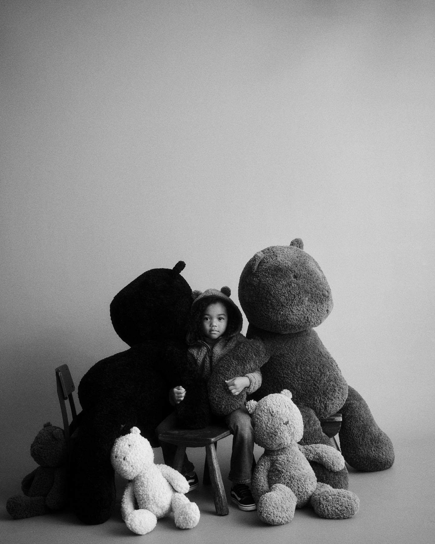 A young child sits on a stool surrounded by large plush teddy bears in a grayscale studio shot, creating a soft and whimsical scene.