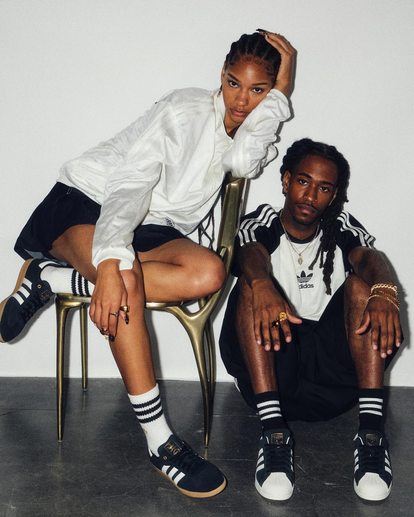 Two fashion models in casual Adidas apparel pose in a photo studio. A woman sits on a gold chair with a man seated next to her.