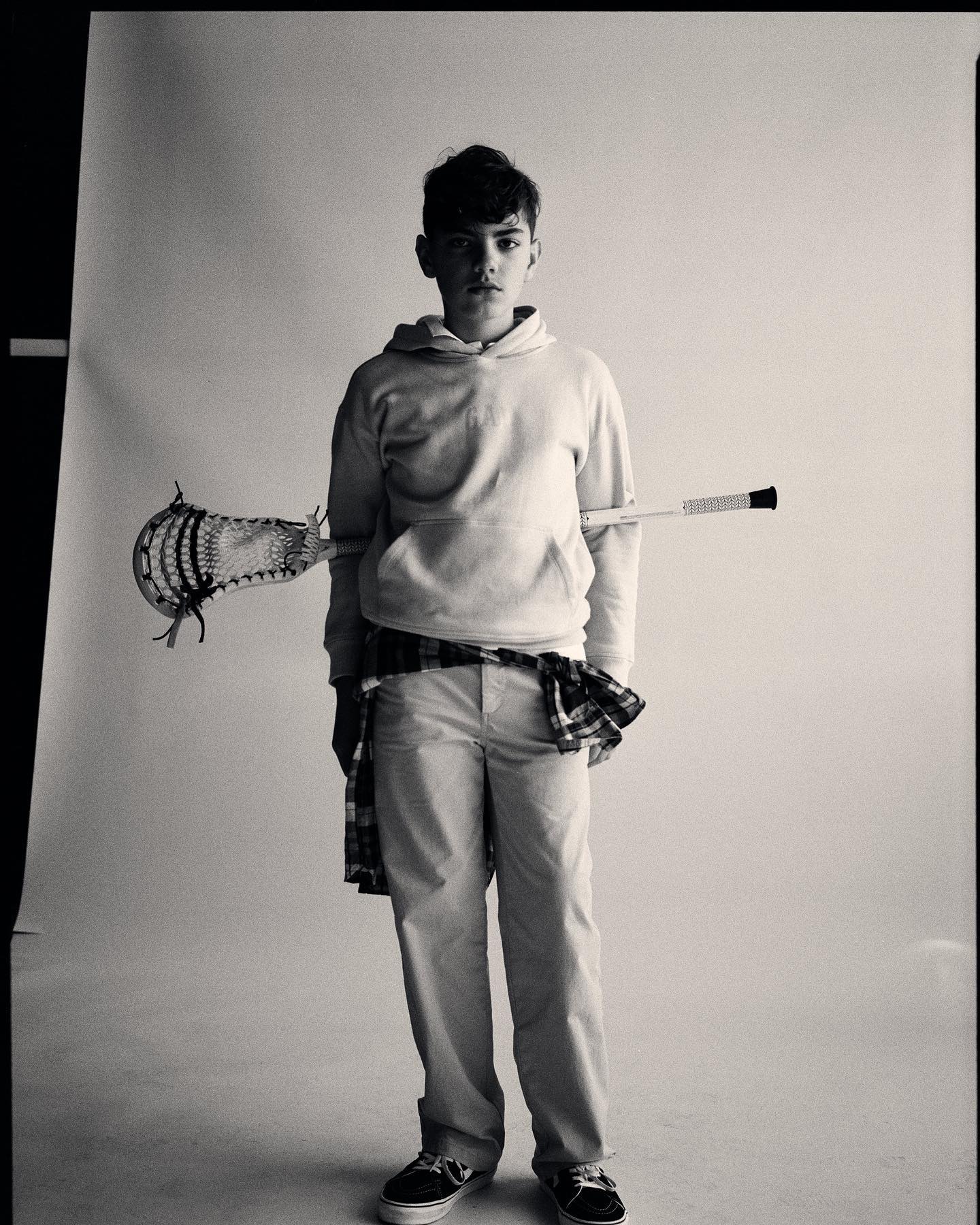 Black and white studio portrait of a young man in a hoodie and slacks, holding a lacrosse stick with a flannel shirt tied around his waist.