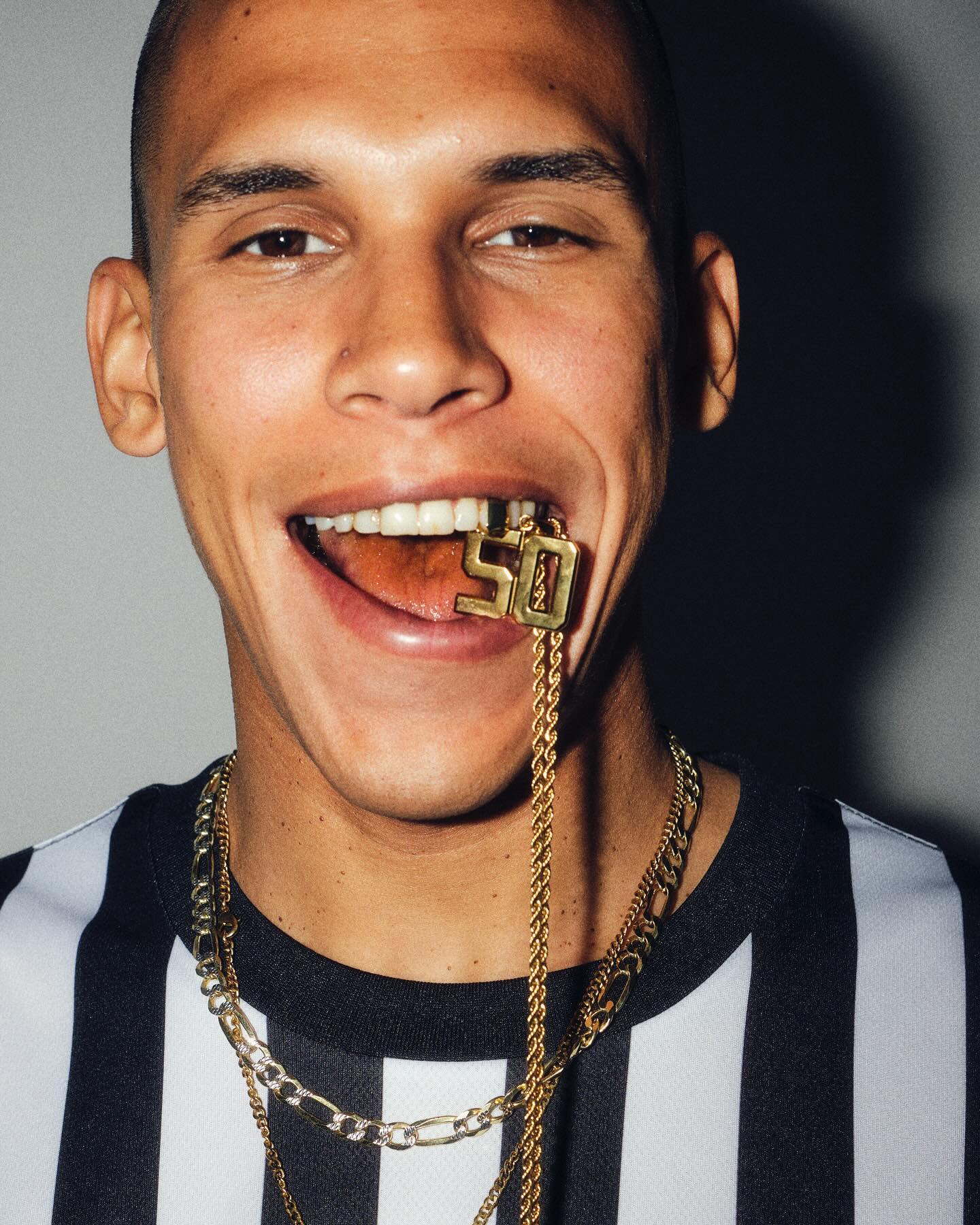 Close-up of adult male with an open mouth wearing a striped shirt and gold chains, one of which has a golden '50' pendant.