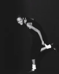 Black and white portrait of a smiling woman adjusting her white socks and dark shoes against a dark background.