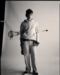 Young boy holding a lacrosse stick behind his back, wearing a hooded sweatshirt and casual trousers against a seamless backdrop