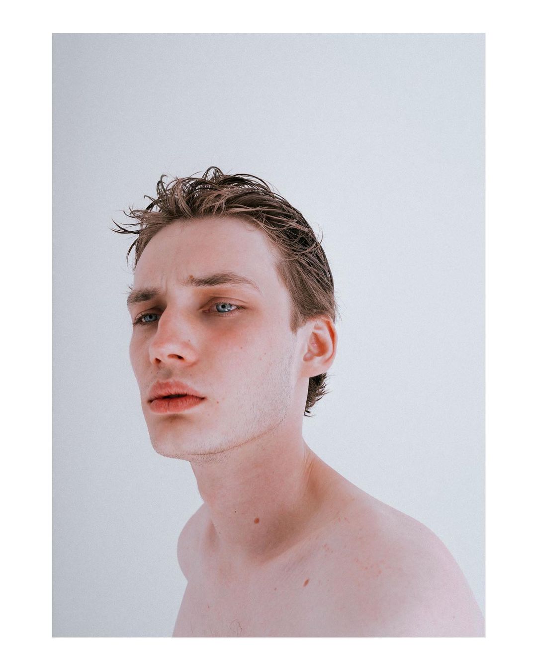 Portrait of a young adult male with wet brown hair, pale skin, and blue eyes, looking off-camera against a bright neutral backdrop in a studio.