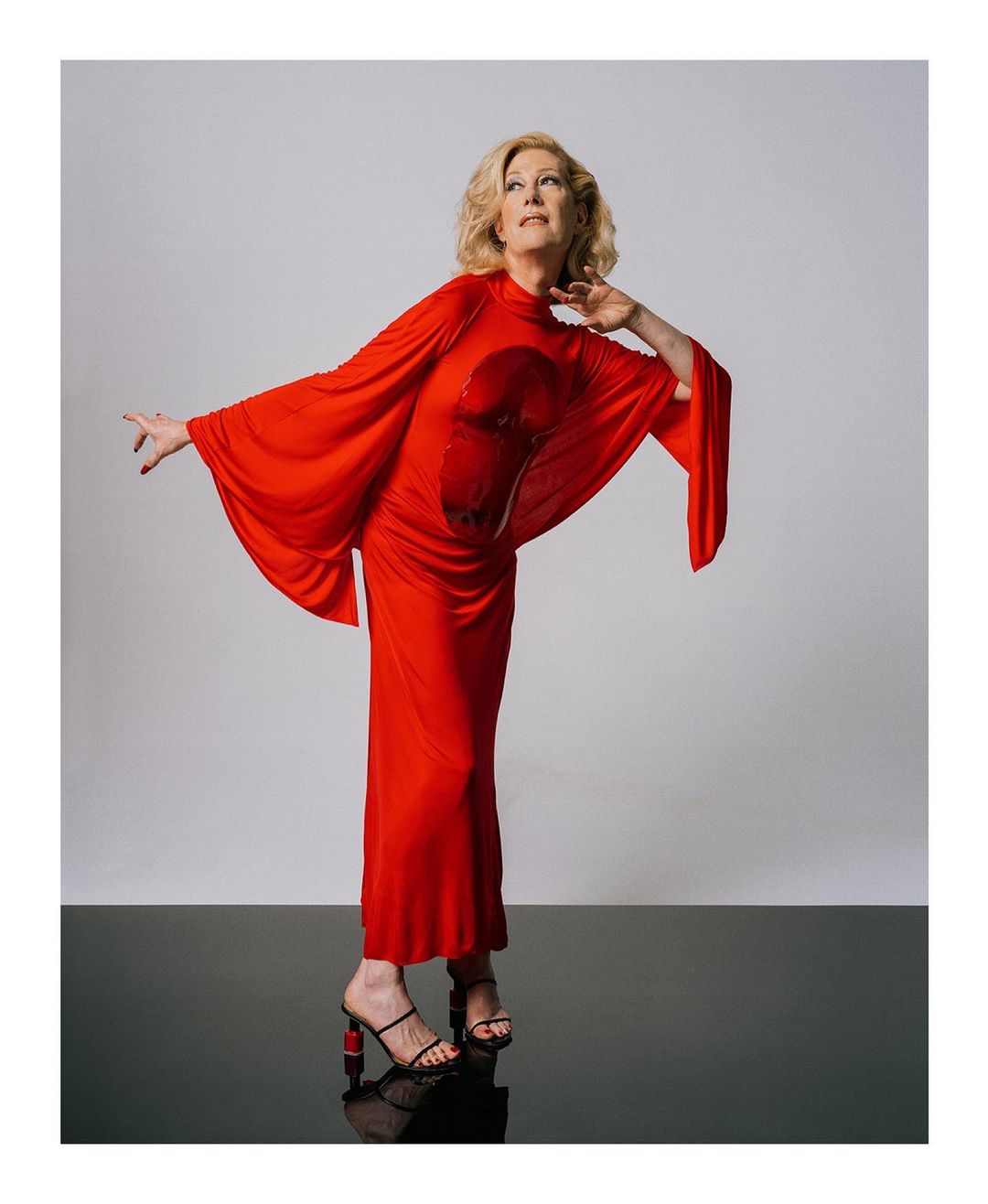 Elegant woman in a red gown poses gracefully in a studio with a smooth reflective floor, showcasing her style and confidence.