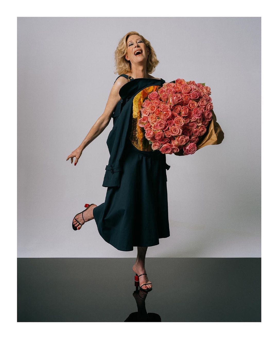 Adult female in a black dress holds a large bouquet of pink roses indoors, smiling and posing against a neutral background with reflection.