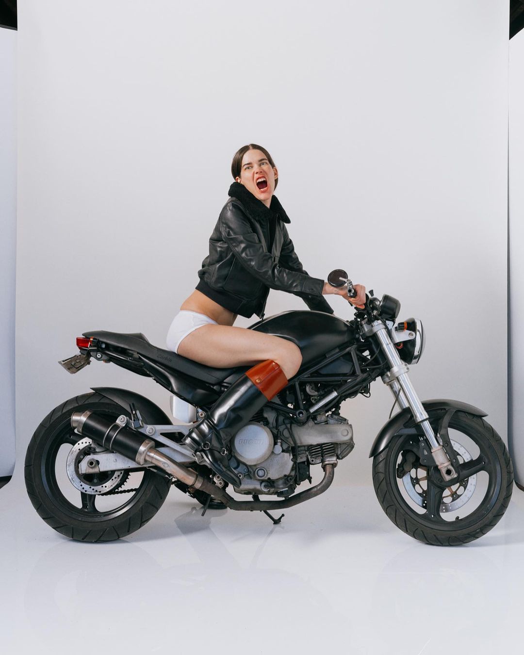 A woman in a leather jacket and white shorts sits astride a black motorcycle in a photo studio against a white backdrop.