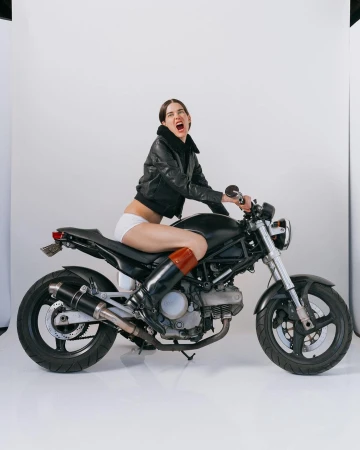A woman in a leather jacket and white shorts sits astride a black motorcycle in a photo studio against a white backdrop.