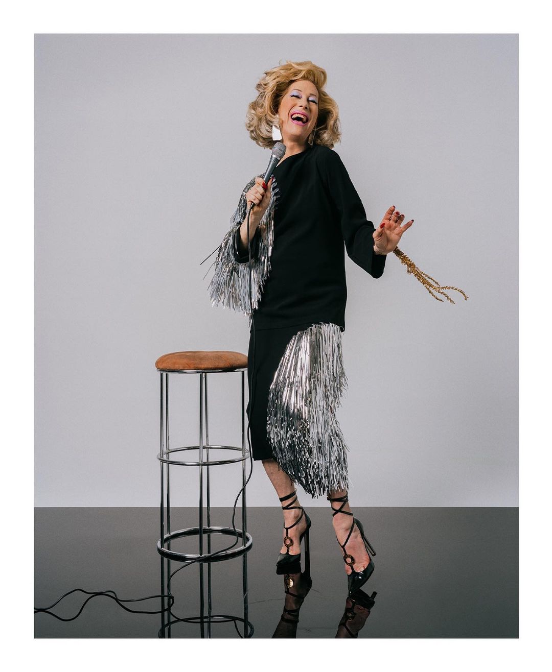 Candid shot of a joyful senior woman singing into a microphone, dressed in a black dress with silver fringe, on black reflective floor.