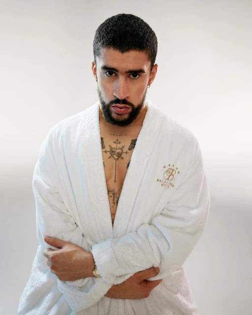 Adult male in a white bathrobe stands against a white background. He has dark hair, a beard, and tattoos on his chest and neck.