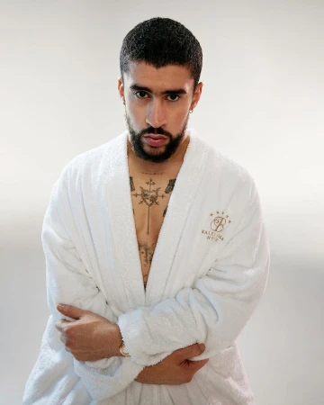Adult male in a white bathrobe stands against a white background. He has dark hair, a beard, and tattoos on his chest and neck.