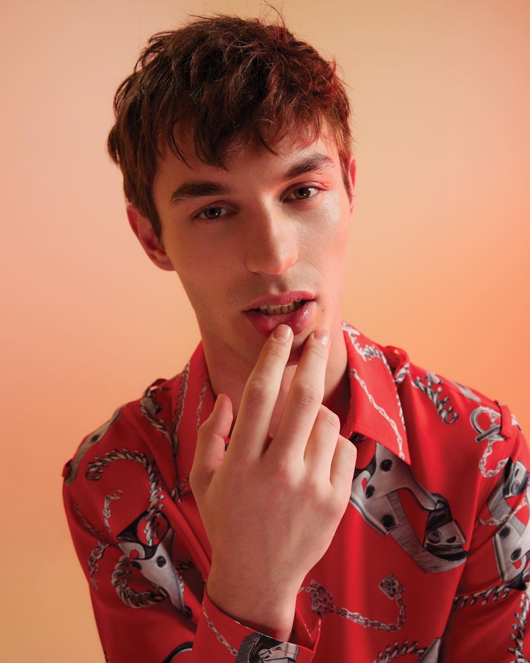 A young adult male model with fair skin poses indoors, wearing a red shirt with chain patterns and pink eyeshadow; close up shot.