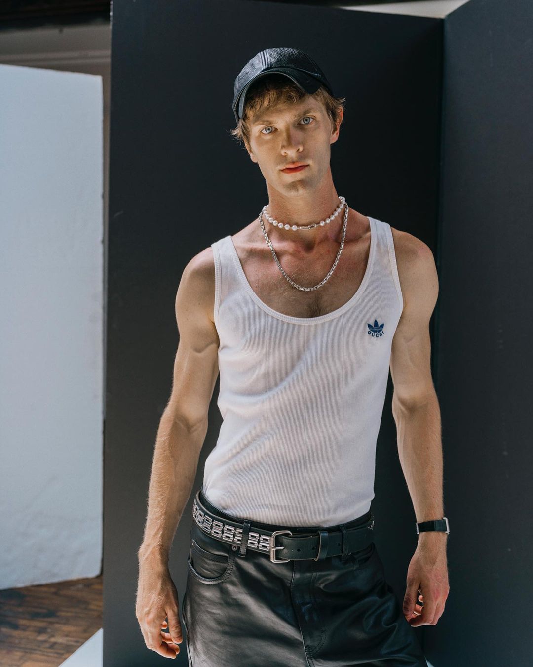 Portrait of a young adult man in a white tank top, leather cap, and black leather skirt posing against a white and black wall indoors.