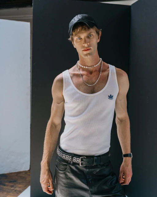 Portrait of a young adult man in a white tank top, leather cap, and black leather skirt posing against a white and black wall indoors.