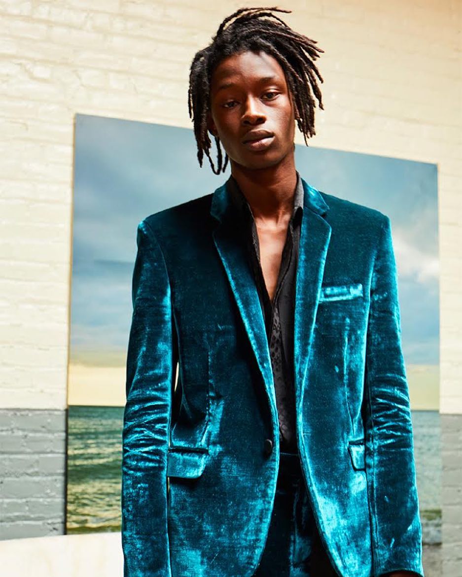 A Black male model wearing a teal velvet blazer and pants poses in front of a painting of a seascape in a studio setting.