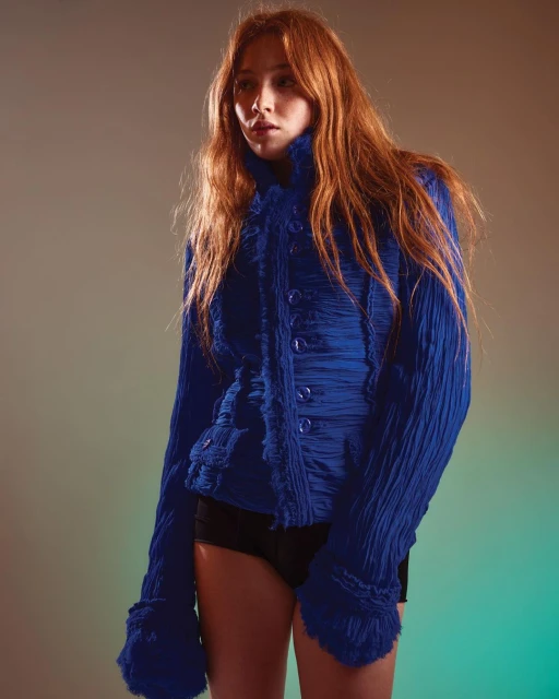 A fashionable young woman with auburn hair poses in a blue textured jacket in a studio setting with soft, diffused lighting.
