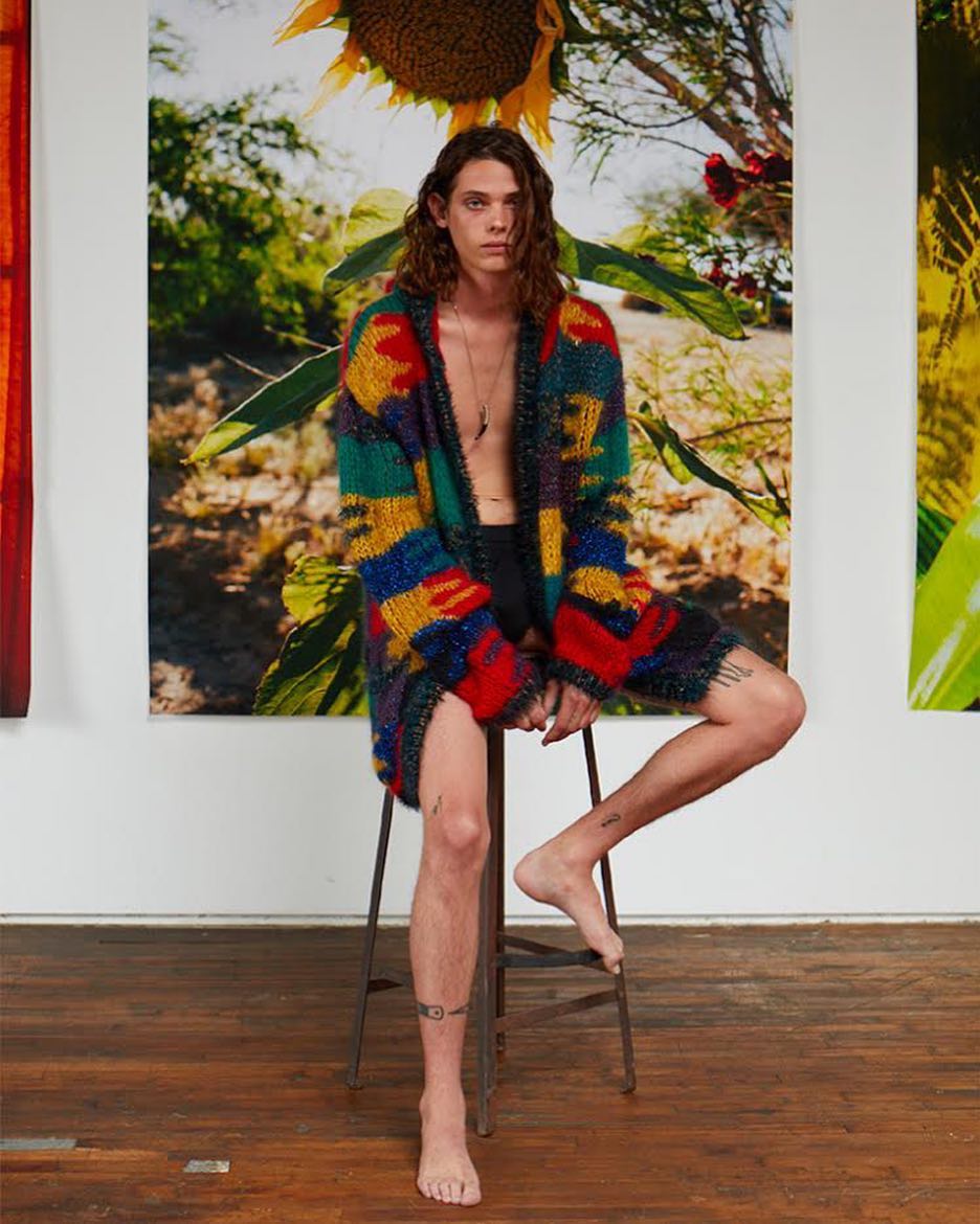 Male model in a colorful cardigan sits on a stool in front of a sunflower photo, showcasing a blend of fashion and art.
