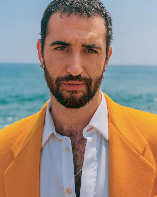 Close-up portrait of a bearded man wearing a gold blazer and white shirt against a blue ocean backdrop, exuding a relaxed and stylish vibe.
