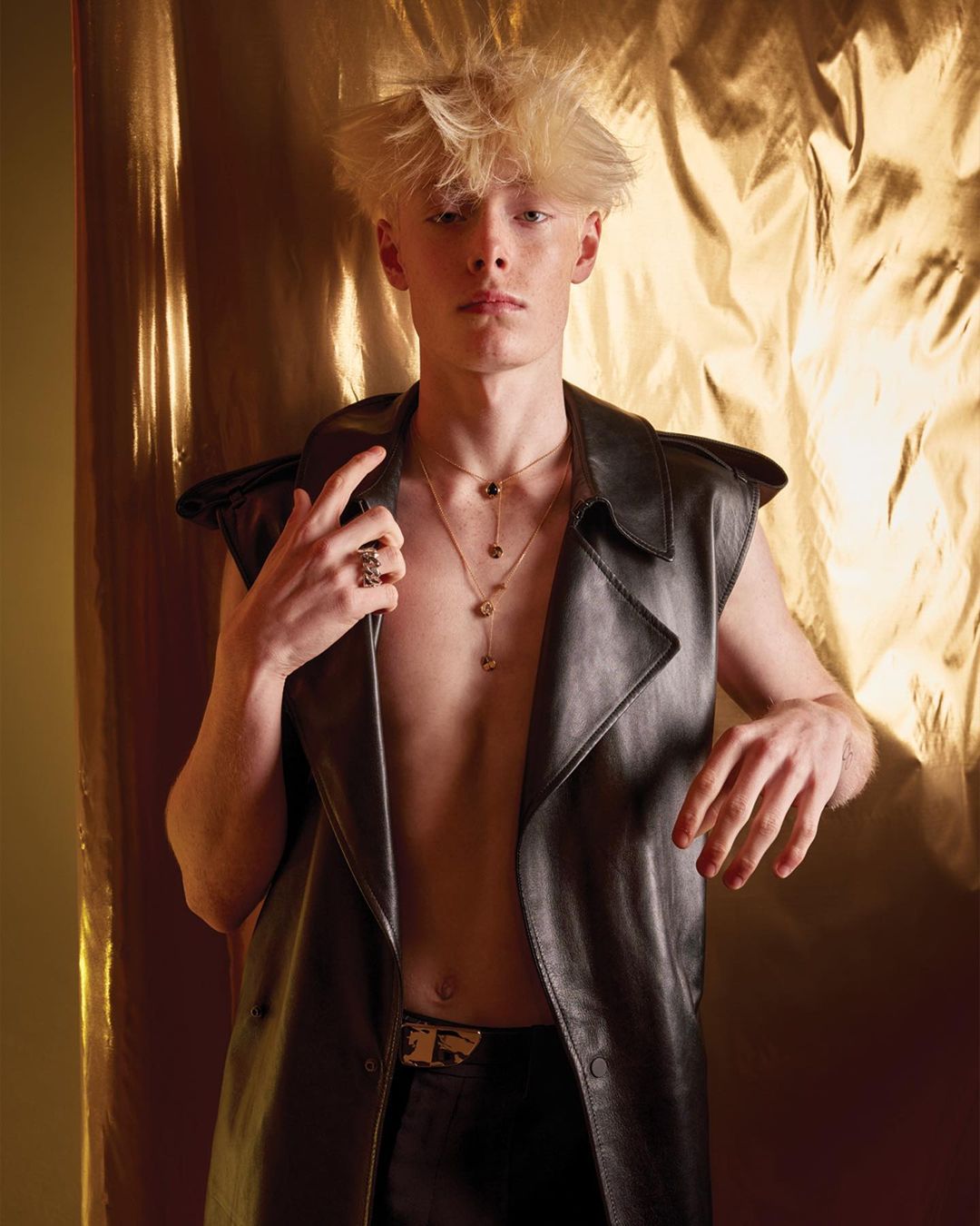 A blond male model in a black leather vest and pants poses in front of a gold, textured backdrop with a serious expression in a studio setting.