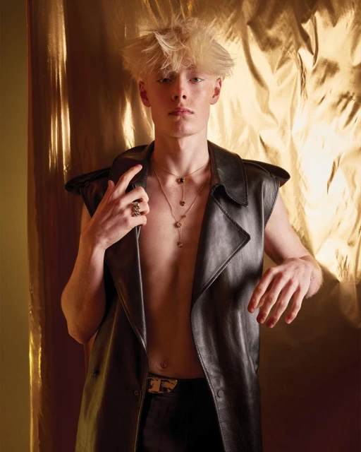 A blond male model in a black leather vest and pants poses in front of a gold, textured backdrop with a serious expression in a studio setting.