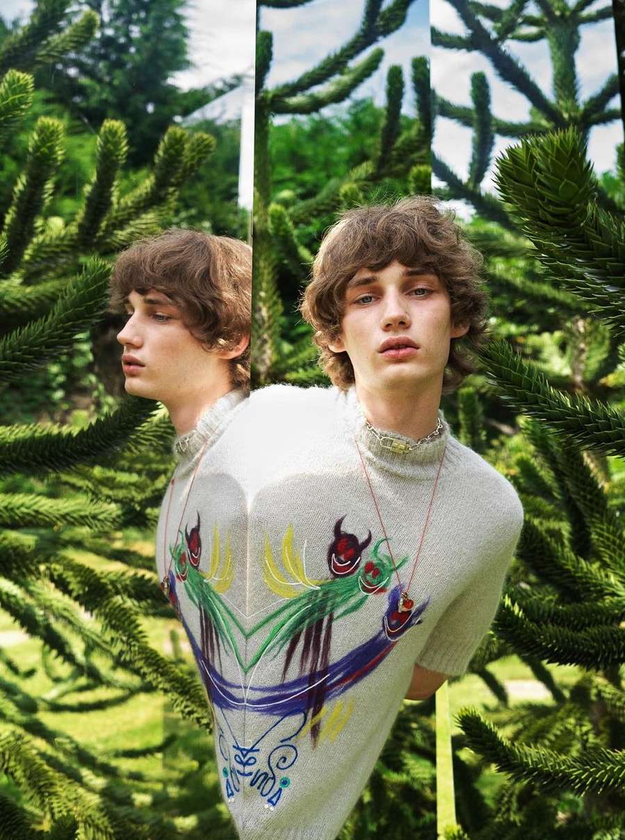 Artistic portrait of a young man reflected in mirrors, framed by lush green foliage in an outdoor setting, showcasing editorial fashion.