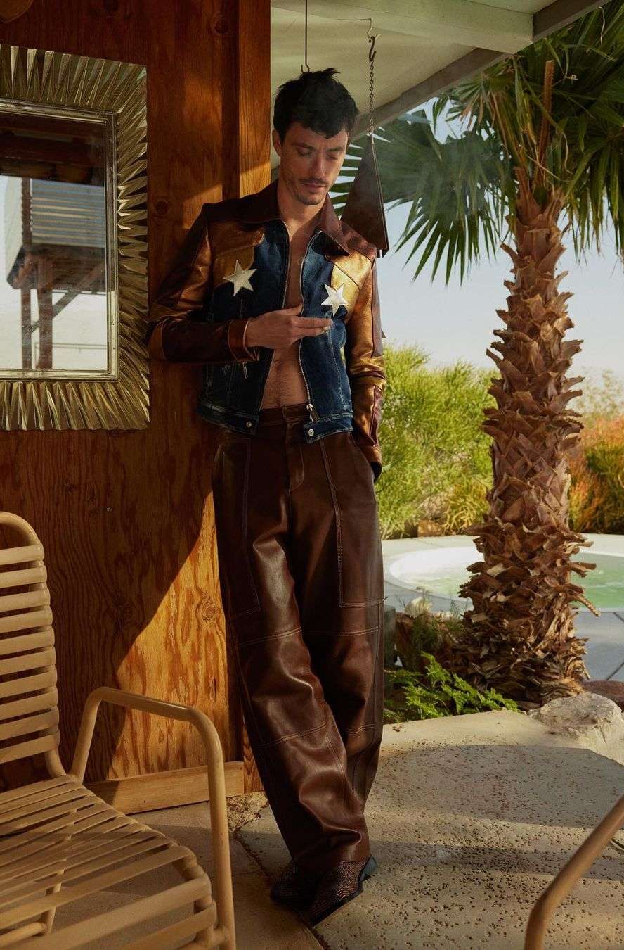 Adult man in an open western-style jacket and brown leather pants stands outdoors, checking his smartphone near palm trees.