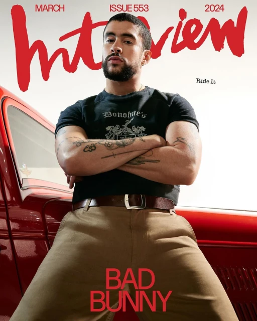 Bad Bunny poses next to red vintage car on magazine cover. Rapper with crossed arms, t-shirt, khaki pants, and a brown leather belt.
