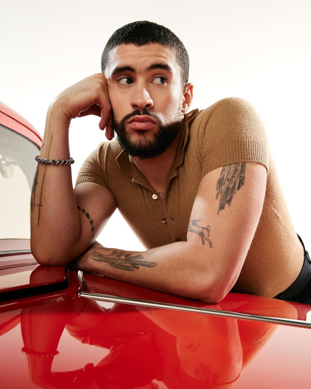 Bad Bunny poses leaning on a classic red car, wearing a polo shirt and bracelet, in a studio shot with a bright white background.