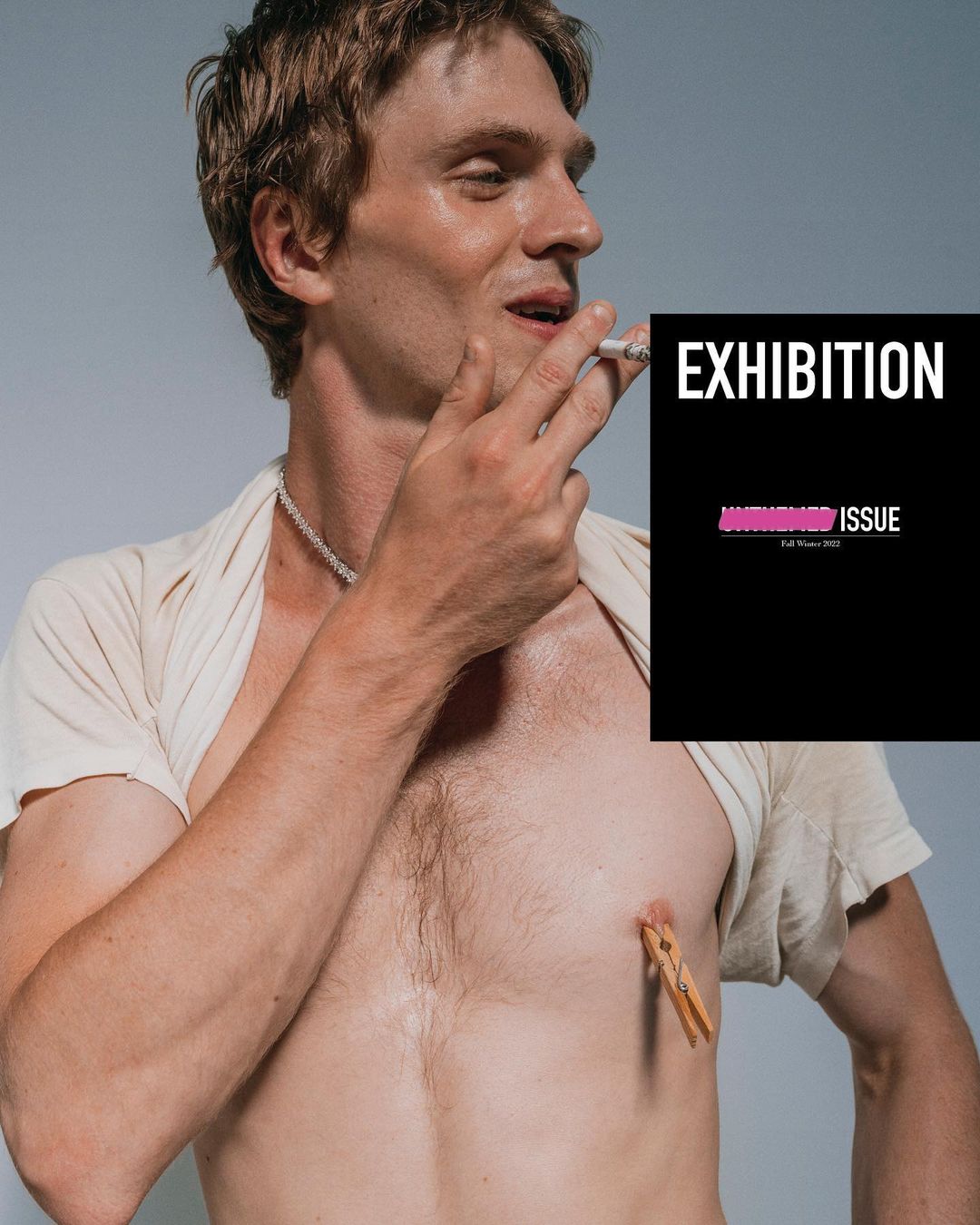 A shirtless adult man poses indoors with a cigarette and a clothespin attached to his chest with a black magazine cover overlay.