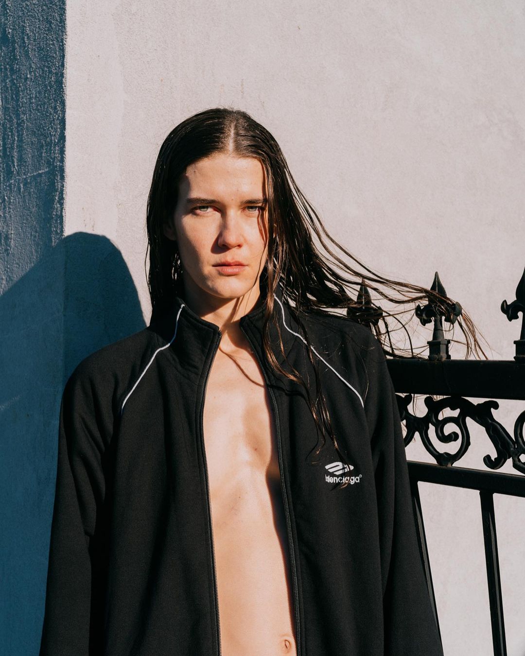 Adult with long hair stands against a white wall in a black jacket, with decorative ironwork to the side, in natural lighting.