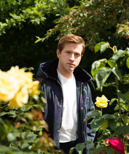 Portrait of a red-haired man wearing a jacket standing among yellow roses and green foliage in an outdoor garden setting on a sunny day.