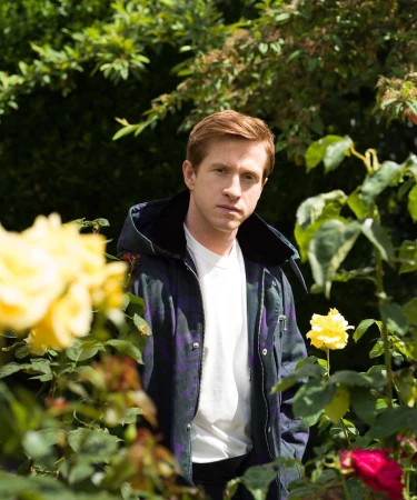 Portrait of a red-haired man wearing a jacket standing among yellow roses and green foliage in an outdoor garden setting on a sunny day.