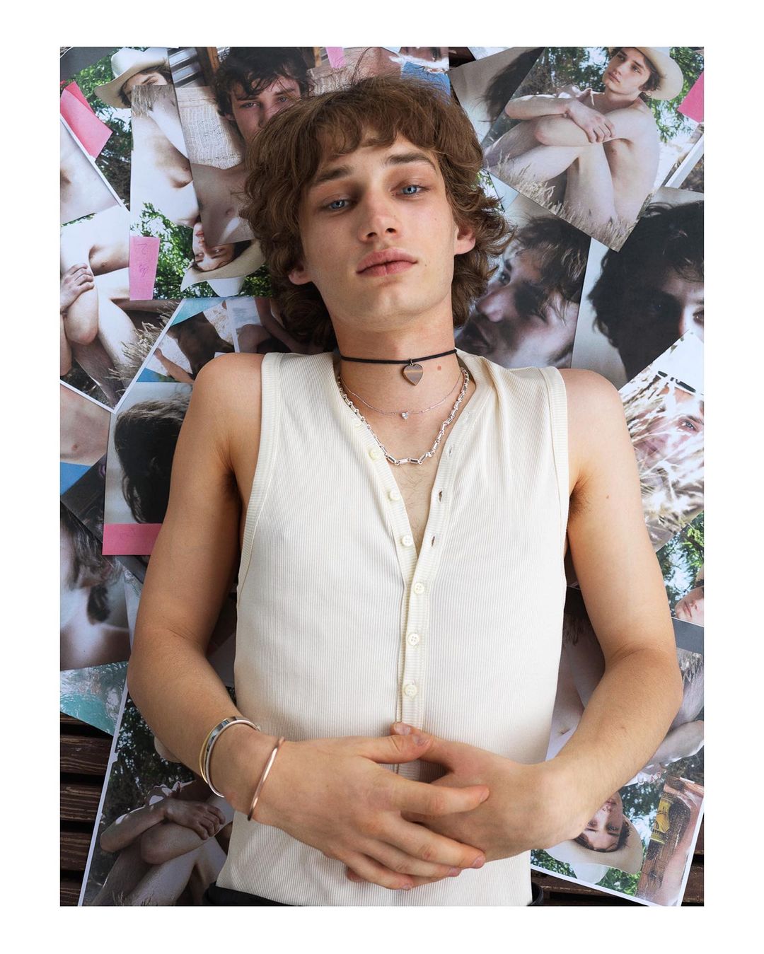 A young adult male model lies on a flat surface surrounded by fashion photography, showcasing a casual white tank top and jewelry.