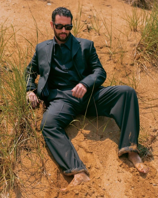 A man in a dark suit sits in the sand on a beach with tall grass wearing sunglasses in an outdoor fashion shot, with a medium shot.
