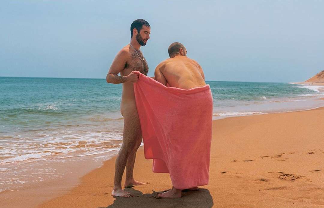 Two adult males on a sandy beach are partly covered by a pink towel, with the ocean and shoreline visible in the background.