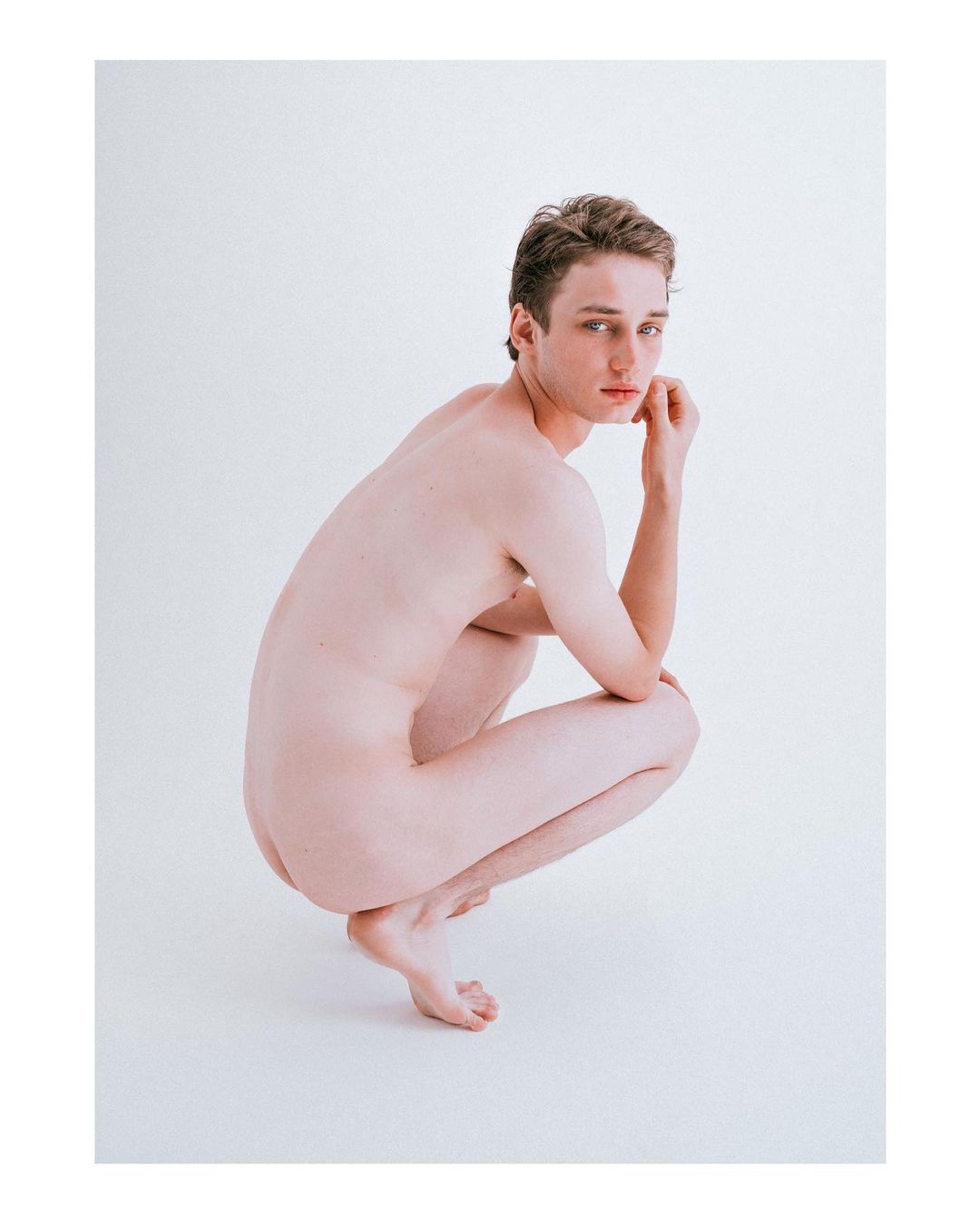 Nude male figure crouching in a white studio environment, looking at the camera with neutral expression, arms resting on legs.